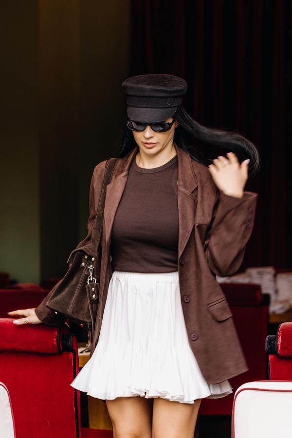 Portrait of a woman in brown blazer, black sailor cap, black sunglasses, white pleated skirt, cafe interior, Paris
